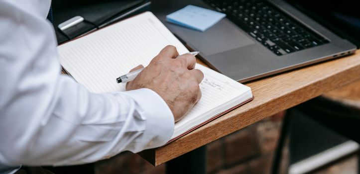 Person taking notes in a notebook next to a laptop while comparing specs