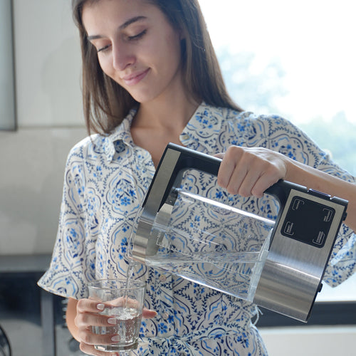Bright lifestyle kitchen scene in soft daylight of a young woman in a blue-print shirt pouring hydrogen water from the silver DR.WATER HydroPitcher into a clear glass she is holding, subject left of center with the pitcher mid-right and ample window light upper-right leaving comfortable negative space for text overlay. No burned-in graphics, suited as a lifestyle hero or pouring-moment block on light email shells.