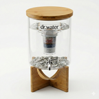Dr.Water TruGlass countertop water filter hero shot on white: a clear glass cylindrical jar with bamboo lid on a crossed bamboo stand, a multi-stage cartridge with red, blue and grey mineral layers hanging inside, and a stainless steel spigot in a bed of grey mineral stones at the base.