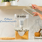 Dr.Water TruGlass dispenser on a white kitchen counter being topped up from a clear glass pitcher held by a hand entering top-right, with a navy handwritten Filters Contaminants callout and curved arrow pointing to the dark filter cartridge inside the tank.