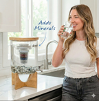 Dr.Water TruGlass lifestyle shot of a smiling brunette woman in a white t-shirt drinking from a clear glass beside the dispenser on a marble counter, with a handwritten Adds Minerals callout in navy and a curved arrow pointing to the mineral bed at the base.