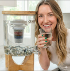 Dr.Water TruGlass lifestyle shot of a smiling brunette woman in a white shirt with green nail polish leaning beside the dispenser holding a clear glass of water, multi-coloured mineral cartridge and pebble bed visible through the glass tank.