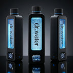 Hero marketing trio of three black rectangular Dr.Water Hydrator Max hydrogen water bottles in a row on a glossy reflective surface against a dark charcoal gradient studio backdrop; the centre bottle has a brightly glowing blue water window while the flanking bottles show subdued lighting variants, with the dr.water wordmark on each bottle window and small touchscreen icons on the base.