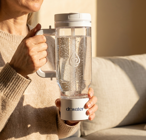 Lifestyle shot of a woman in a cream knit sweater holding the white HydroTumbler with both hands while seated on a warm beige sofa, soft window light around her, torso and face cropped at top so the bottle stays centered with cozy negative space below. No overlays or burned-in text. Used as a lifestyle hero or storytelling block in welcome emails; warm tones pair naturally with cream and beige email backgrounds.