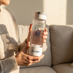 Woman in cream sweater holding the white-cap Dr.Water HydroDaily 13oz hydrogen bottle on a beige sofa in soft warm daylight.