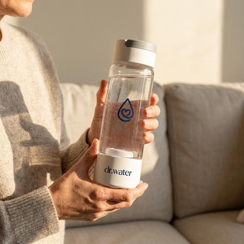 Woman in cream sweater holding the white-cap Dr.Water HydroDaily 13oz hydrogen bottle on a beige sofa in soft warm daylight.