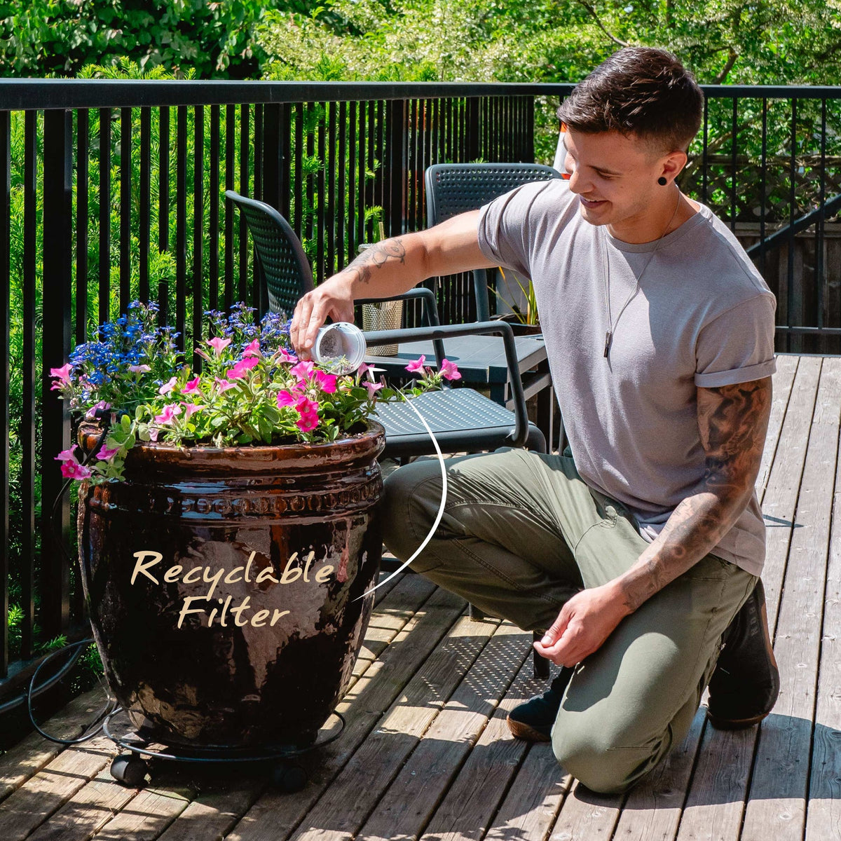 Dr.Water TruGlass lifestyle photo of a tattooed man in a grey t-shirt and olive pants kneeling on a sunlit wooden deck, watering pink and blue flowers from a clear glass beside a black wheeled planter, with a white handwritten Recyclable Filter callout pointing to the planter.
