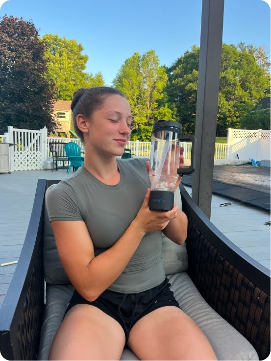 Lifestyle UGC photo of a smiling woman in an olive green top seated on a black wicker chair on a backyard deck, holding a black Dr.Water HydroTumbler hydrogen water bottle in both hands at chest level with greenery and a white fence behind her.