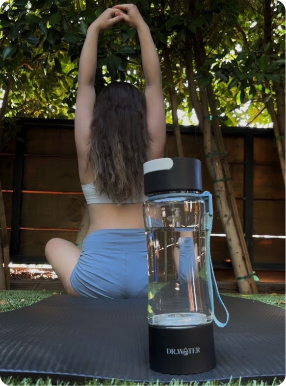 Lifestyle photo of a woman in a light bra and grey shorts kneeling with arms stretched overhead on a black yoga mat in a backyard, with a clear Dr.Water hydrogen water bottle on a black DR.WATER base in the foreground and bamboo fencing and greenery behind.