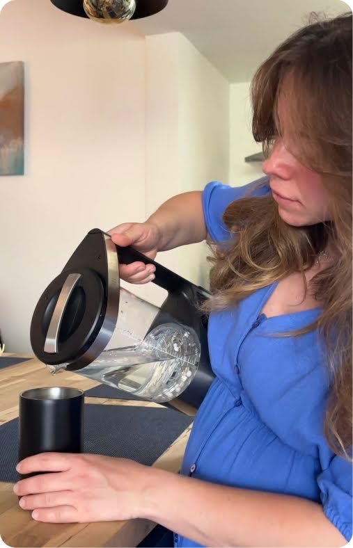 Lifestyle UGC photo of a woman in a blue blouse at a wood dining table pouring water from a black-lidded Dr.Water glass kettle into a black tumbler base, with a framed painting on a white wall behind her in soft indoor light.