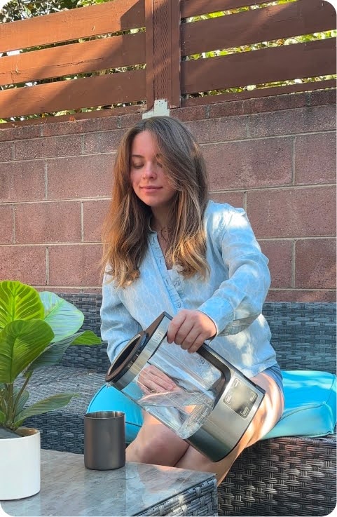 Lifestyle UGC photo of a woman in a light blue shirt seated on an outdoor wicker couch pouring filtered water from a clear Dr.Water glass kettle into a black tumbler base on a marble side table, with a potted plant and brick fence in warm golden-hour light.